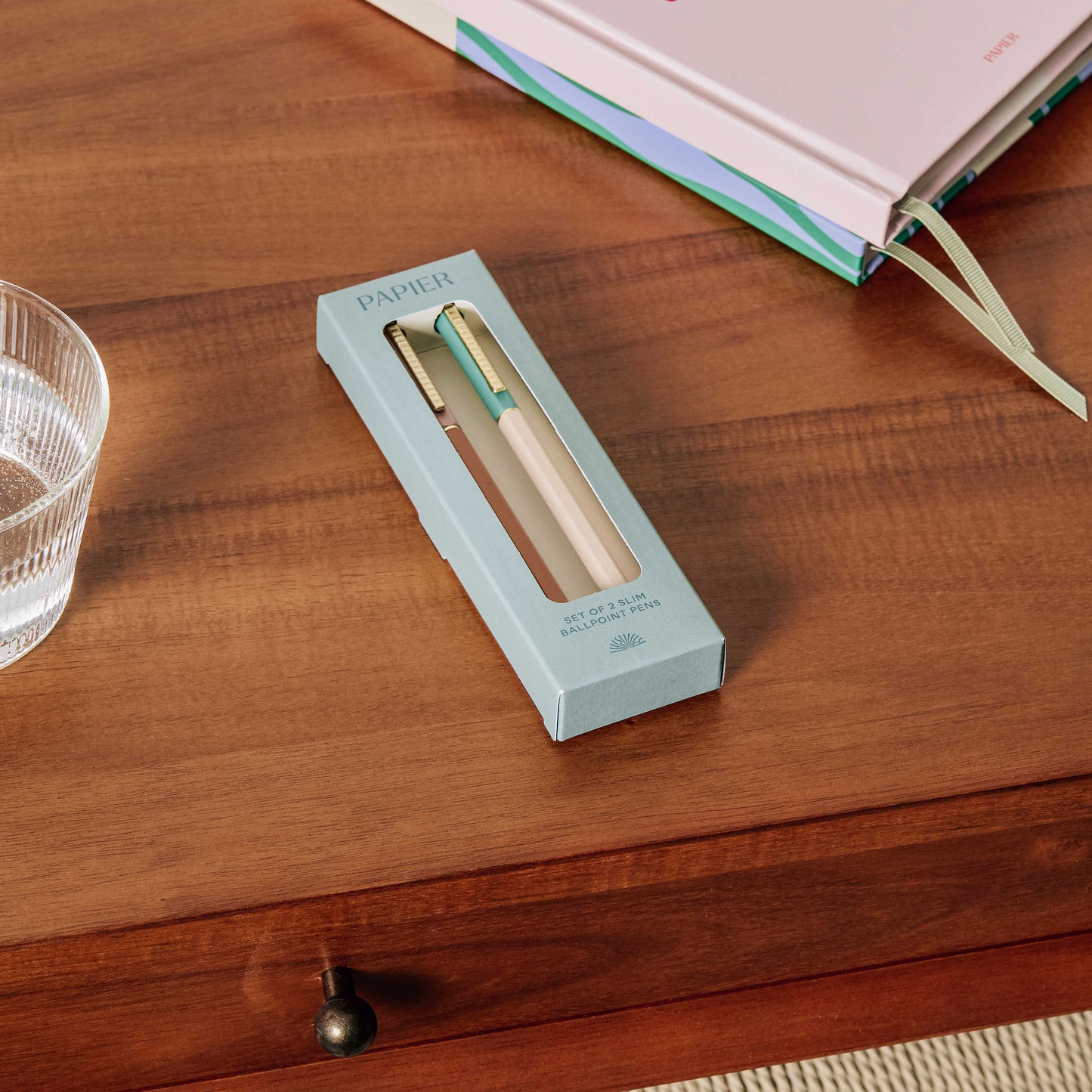 Papier pen set in a box on a wooden surface with a glass of water and a book.