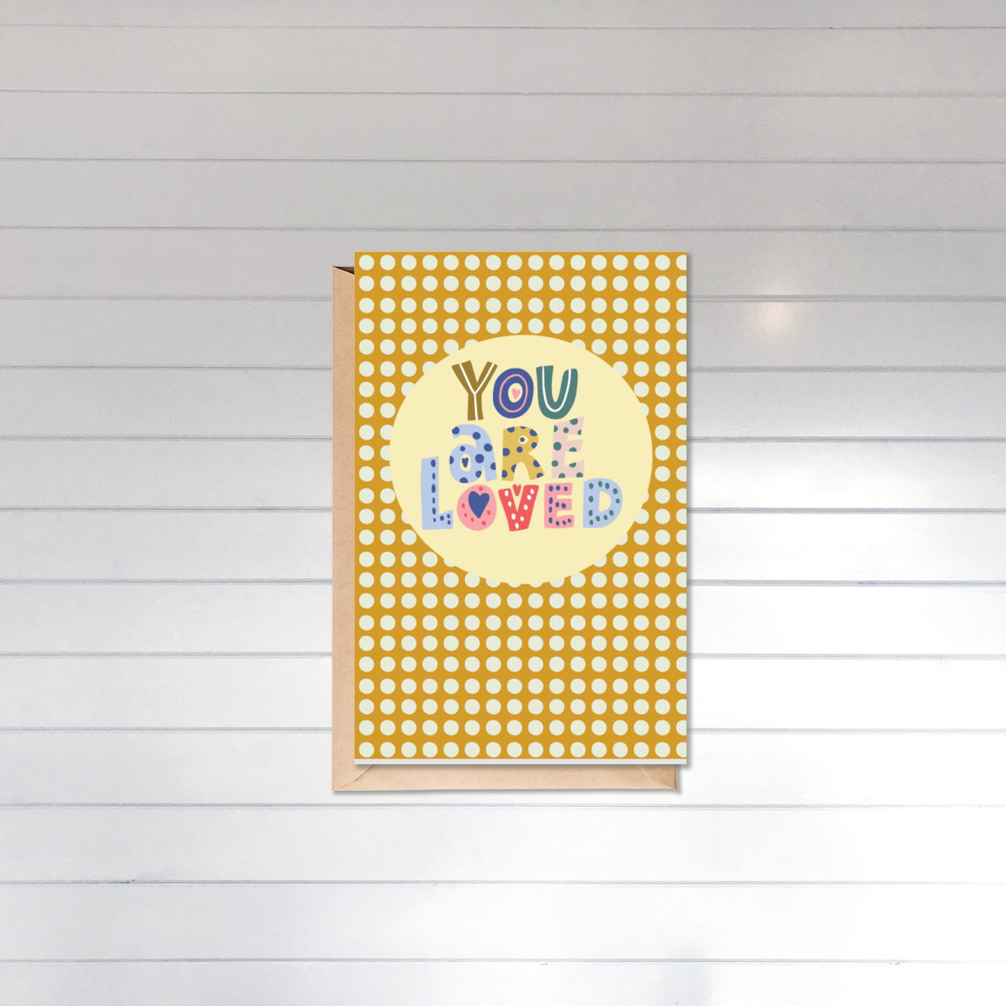 Greeting card with 'You are loved' text on a yellow polka dot cover against a white wooden background